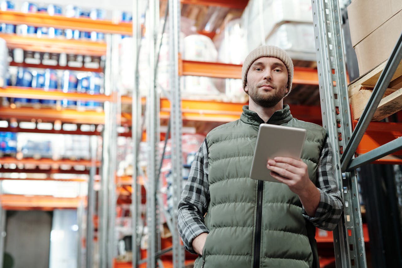 Young male warehouse manager in a beanie holding a tablet in a storeroom.